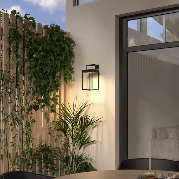 Moderne schwarze Außenwandleuchte mit rechteckigem Rahmen und warmem Licht an einer hellen Hauswand neben einem Holzzaun mit Grünpflanzen.