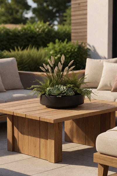 Holz-Loungetisch im Außenbereich mit einer Pflanzschale aus Sukkulenten, Farnen und Ziergräsern, umgeben von hellen Polstermöbeln.