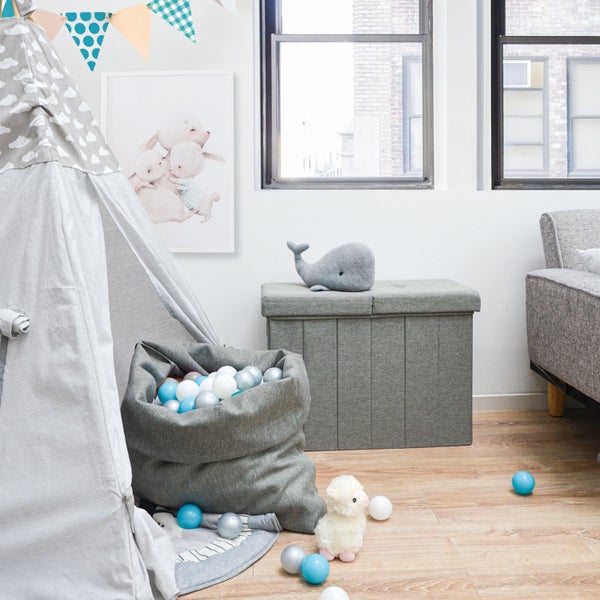 Helles Kinderzimmer mit einer grauen Sitztruhe aus Stoff, einem großen Spielzeugsack voller Bälle, einem Tipi-Zelt und Boden in Holzoptik.