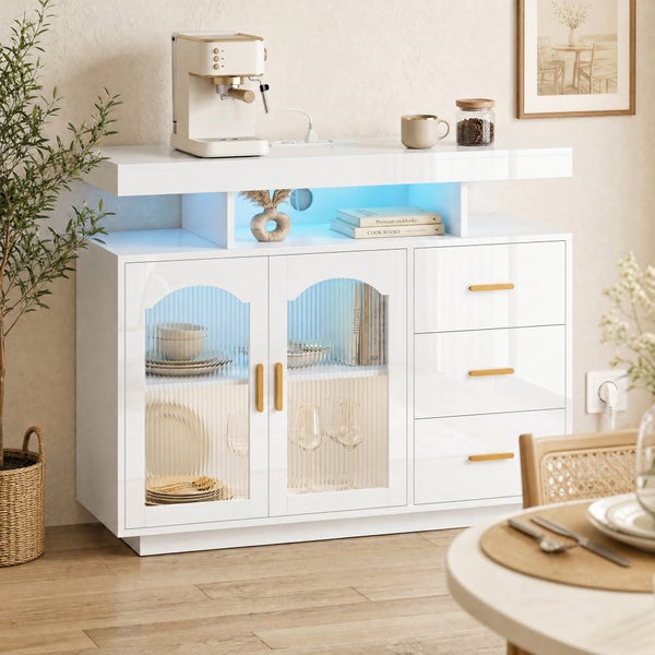 Modernes weißes Sideboard mit blauer Beleuchtung, geriffelten Glastüren und Goldgriffen in einem hellen Raum mit Pflanze und Esszimmermöbeln.