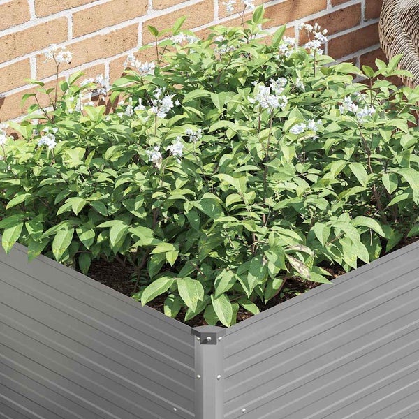 Hochbeet aus grauem Metall mit gewellter Oberfläche, bepflanzt mit grünem Laub und weißen Blüten vor einer Ziegelwand.