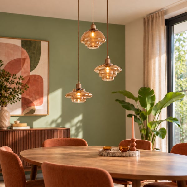 Drei hängende Pendelleuchten mit gewölbten Glasschirmen über einem runden Esstisch in einem modernen Esszimmer mit Wandbild und Pflanzen.