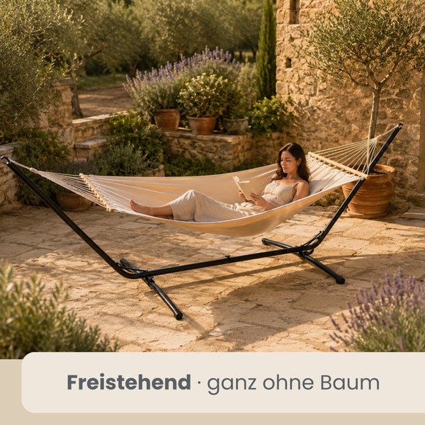 Freistehende Hängematte mit Metallgestell auf einer Steinterrasse im Garten, in der eine Frau ein Buch liest.