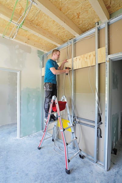 Mann arbeitet mit Bohrmaschine auf einer Leiter in einem Rohbau mit Trockenbauwänden.