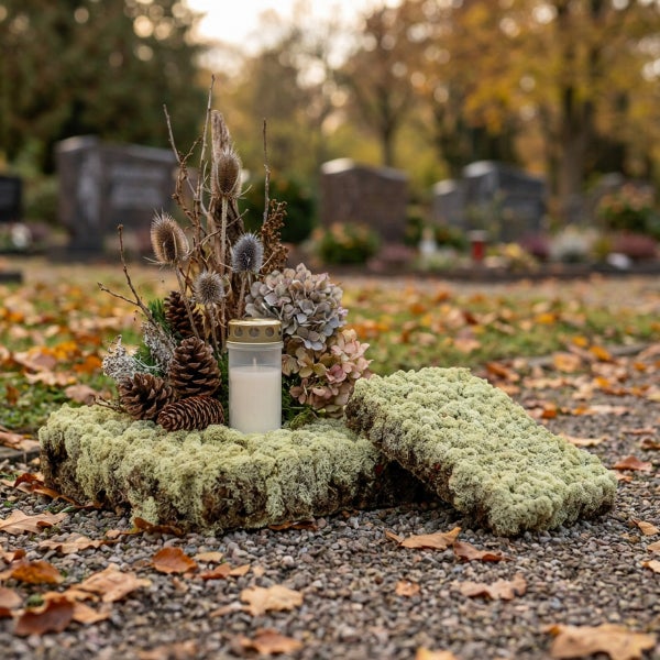 Grabgesteck mit Moos, Tannenzapfen, Trockenblumen und Grablicht auf einem Friedhof, herbstliche Dekoration für eine würdige Grabgestaltung.