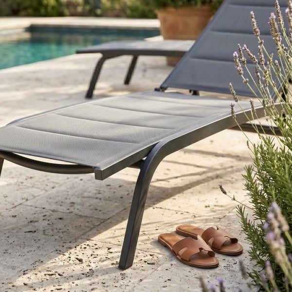 Moderne Gartenliege mit grauem Textilgewebe und Aluminiumgestell auf einer Steinterrasse am Pool.