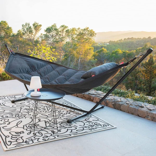 Szene mit einer grauen Hängematte mit Kissen auf einem schwarzen Metallgestell, einem schwarzen Beistelltisch mit Lampe und einem gemusterten Teppich im Außenbereich