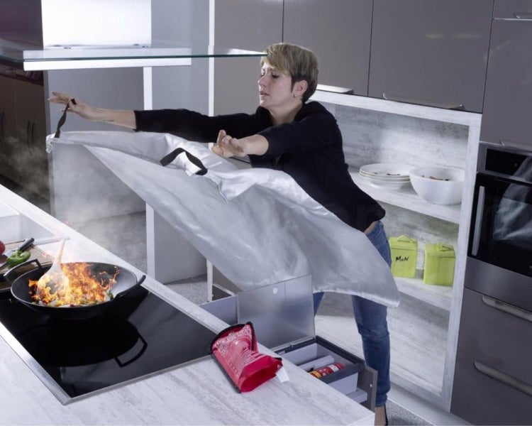 Eine Person breitet eine Löschdecke über eine brennende Pfanne auf einem Induktionskochfeld in einer modernen Küche aus.