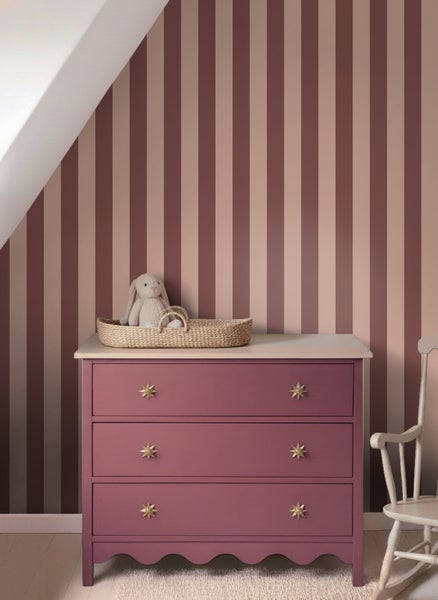 Gestreifte Vliestapete in Altrosa und Beige hinter einer lila Kommode in einem hellen Kinderzimmer.