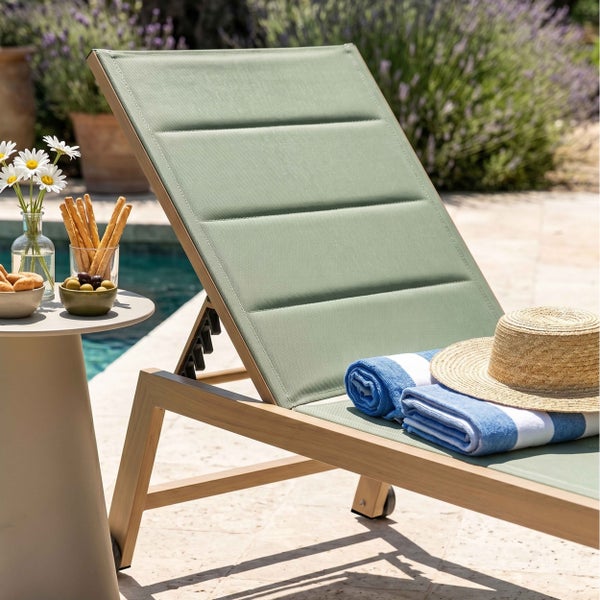 Gartenliege mit hellem Holzgestell und grüner gepolsterter Bespannung am Pool mit Beistelltisch, Handtüchern und Strohhut.