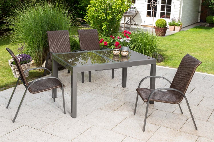 Gartentisch mit Glasplatte und vier Geflechtstühlen auf einer gepflasterten Terrasse in einem Garten mit Gartenhaus.