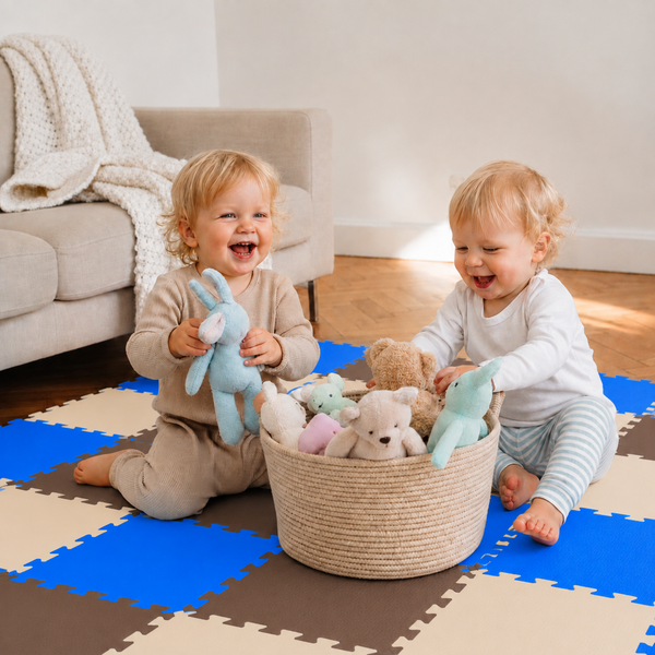 Zwei Kleinkinder spielen auf einer Puzzlematte aus Schaumstoff mit einem Spielzeugkorb voller Stofftiere in einem hellen Wohnzimmer.