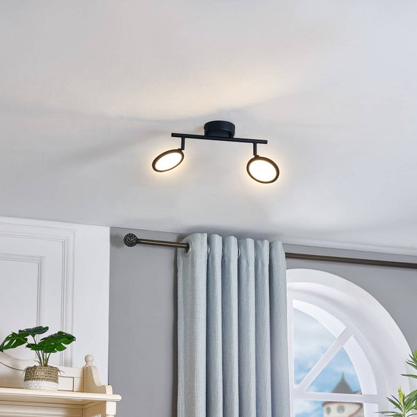 Moderne schwarze Deckenleuchte mit zwei ringförmigen LED-Spots an einer weißen Decke in einem hellen Raum mit grauen Vorhängen und einem Fenster.