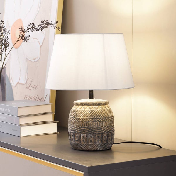 Eingeschaltete Tischleuchte mit strukturiertem Keramikfuß im Ethno-Stil und weißem Textilschirm auf einem Sideboard neben Büchern.