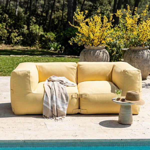 Modulares Outdoor-Sofa in Gelb auf einer sonnigen Terrasse am Pool, mit Beistelltisch, Hut und dekorativen Pflanzen.