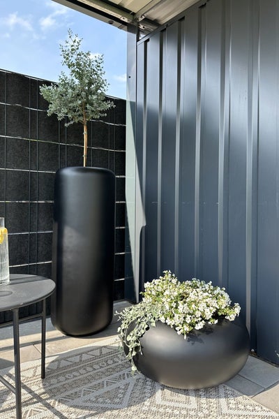 Moderne Terrasse mit einem hohen zylindrischen schwarzen Pflanzgefäß und einer flachen, runden Pflanzschale mit weißen Blumen auf einem Teppich.
