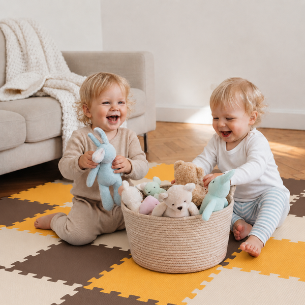 Zwei kleine Kinder spielen lachend mit Stofftieren aus einem runden geflochtenen Korb auf einem gepolsterten Spielboden im Wohnzimmer.