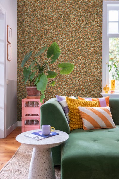 Wohnzimmer mit Leopardenmuster-Tapete, grünem Samtsofa mit bunten Kissen und einer großen Zimmerpflanze auf rosa Kisten.