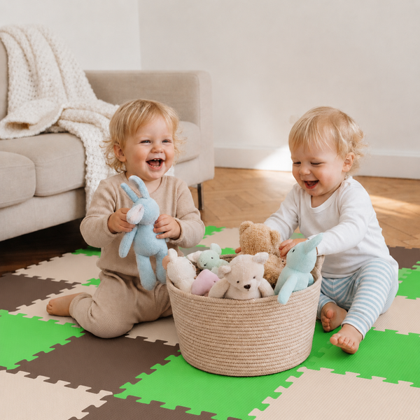 Zwei Kleinkinder spielen lachend auf einer Puzzlematte aus Schaumstoff mit einem geflochtenen Korb voller Kuscheltiere in einem hellen Wohnzimmer.