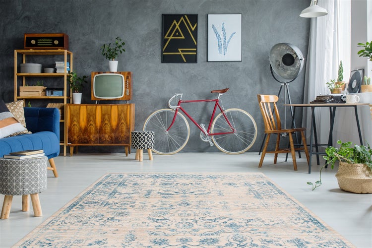 Wohnzimmer im Vintage-Stil mit Teppich mit Orientmuster, blauem Sofa, Holzkommode, Fahrrad an der grauen Wand und dekorativen Zimmerpflanzen.