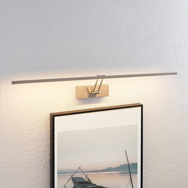 Bilderleuchte aus Metall in Messing-Optik, schlankes Design, über einem Bilderrahmen an einer strukturierten Wand montiert, helles Licht.