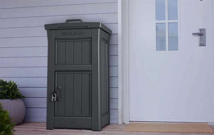 Dunkelgraue Paketbox aus Kunststoff mit Vorhängeschloss neben einer weißen Haustür und blauer Hausfassade.