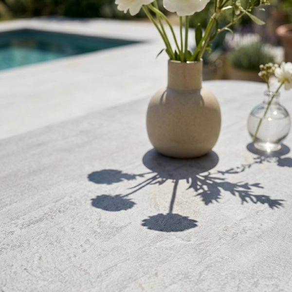 Terrassentisch in Steinoptik im Außenbereich am Pool mit einer Keramikvase und Blumen, die einen Schatten auf die Platte werfen.