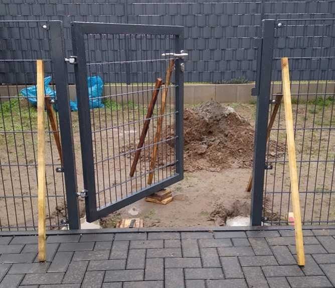 Montage eines anthrazitfarbenen Doppelstabmatten-Gartentors zwischen zwei Zaunpfosten mit Holzstützen auf einer Baustelle im Garten.