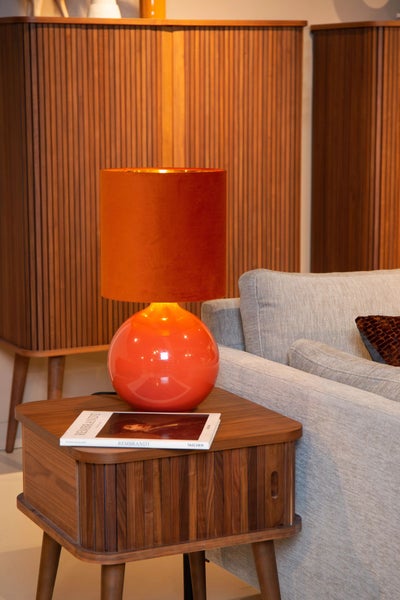 Dekoratives Wohnzimmerambiente mit einer orangefarbenen Tischleuchte, einem Holztisch, einem Sofa und einem Schrank.