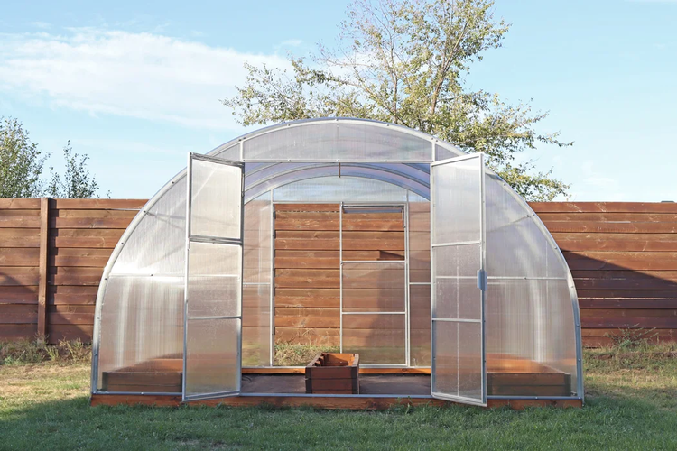 Rundbogen-Gewächshaus aus Polycarbonat mit geöffneten Doppeltüren auf einem Holzsockel im Garten.