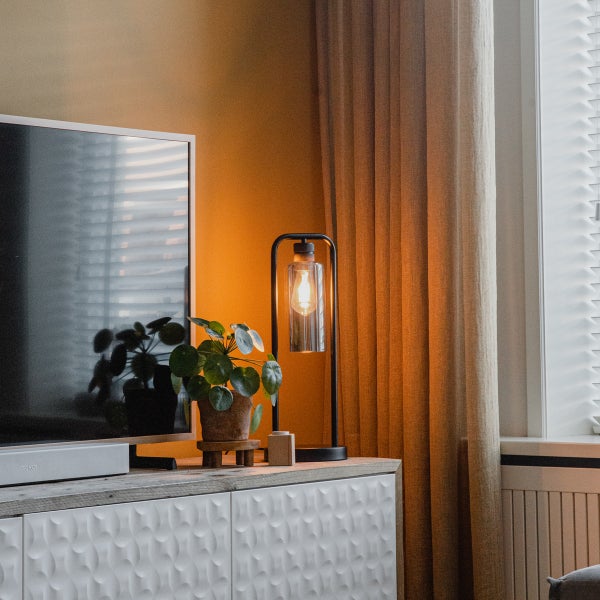 Moderne Wohnzimmerszene mit industrieller Tischleuchte mit Filament-Glühbirne, Zimmerpflanze und Fernseher auf einem strukturierten Sideboard.