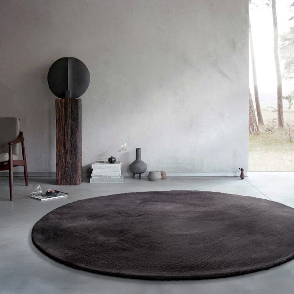 Ein großer runder dunkelgrauer Hochflor-Teppich in einem minimalistischen Raum mit Betonboden und moderner Dekoration.