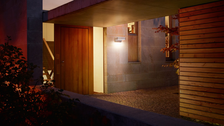 Moderne Außenwandleuchte beleuchtet einen Hauseingang mit Steinwand und Holztür bei Nacht.