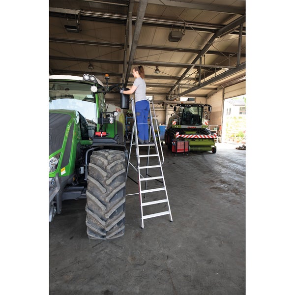 Frau auf einer Aluminium-Stufenleiter bei der Reinigung eines Traktors in einer Werkstatt.