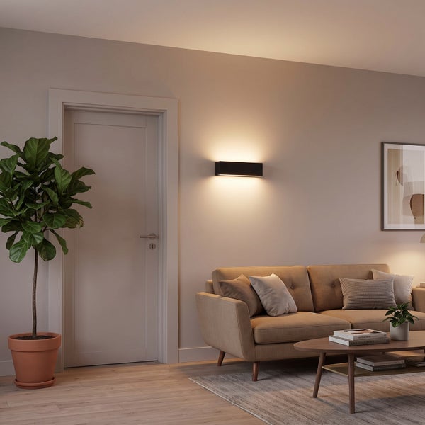 Moderne Wandleuchte in einem wohnlichen Wohnzimmer mit Sofa, Zimmerpflanze und Couchtisch bei warmem Lichtschein.