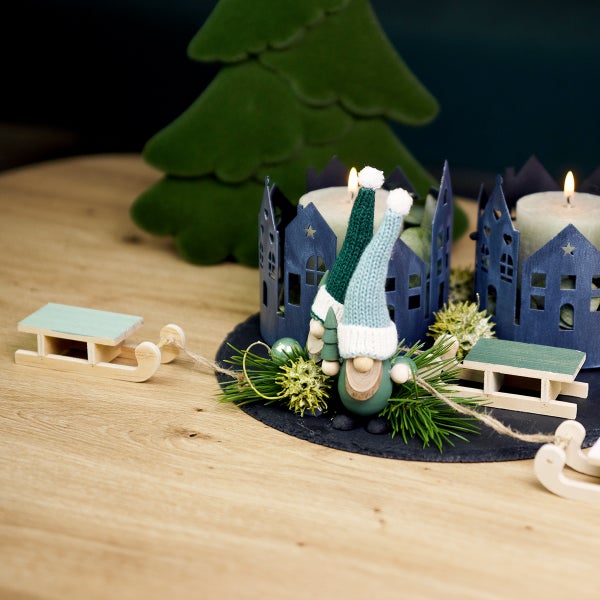 Weihnachtsdekoration mit Kerzen, Häusern, Wichteln und Schlitten auf einem Tisch.