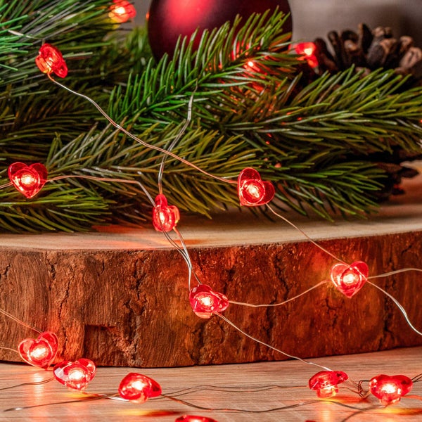 LED-Lichterkette mit roten Herzen auf einem Holzblock neben Tannenzweigen.