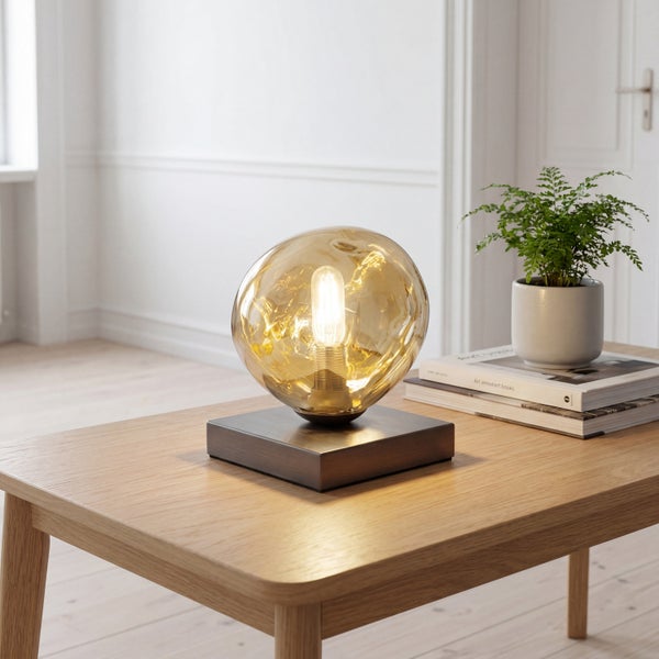 Moderne Tischleuchte mit bernsteinfarbenem Glasschirm auf quadratischem Metallsockel auf einem Holztisch in hellem Raum.