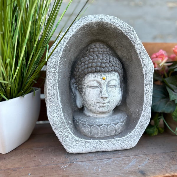 Gartendeko Buddha-Kopf in einer steinähnlichen Nische auf einem Holzregal neben Pflanzen.