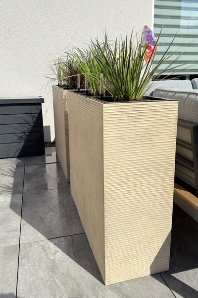 Hohe rechteckige Pflanzgefäße mit horizontaler Rillenstruktur auf einer Terrasse mit Fliesen, bepflanzt mit Ziergras neben einem Gartensofa.