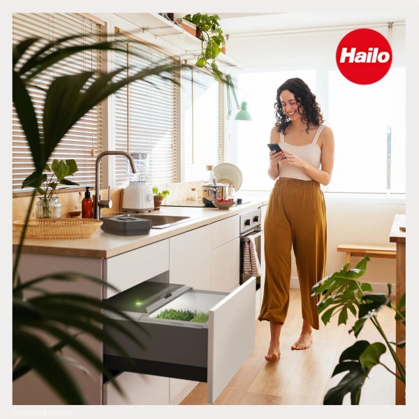 Eine Frau in einer hellen Küche steht neben einem offenen Auszug mit dem Hailo Microgarden zur Aufzucht von Kräutern. Hailo Logo.
