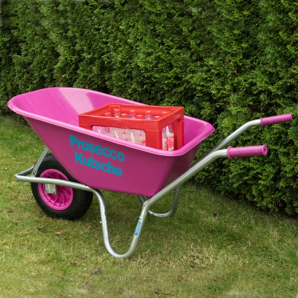 Eine Schubkarre mit der Aufschrift Prosecco Kutsche steht auf einem Rasen vor einer Hecke und ist mit einem Getränkekasten beladen.
