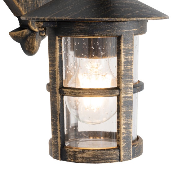 Außenwandleuchte im Laternen-Stil aus Metall in Gold-Antik-Optik mit klarem Bläschenglas und eingeschalteter Lichtquelle.