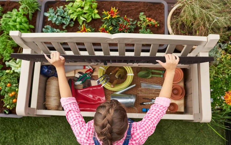 Eine Person öffnet eine Gartenbank mit Stauraum für Gartenwerkzeuge wie Schlauch, Töpfe und Schaufeln inmitten eines Gartens mit Pflanzenbeeten.