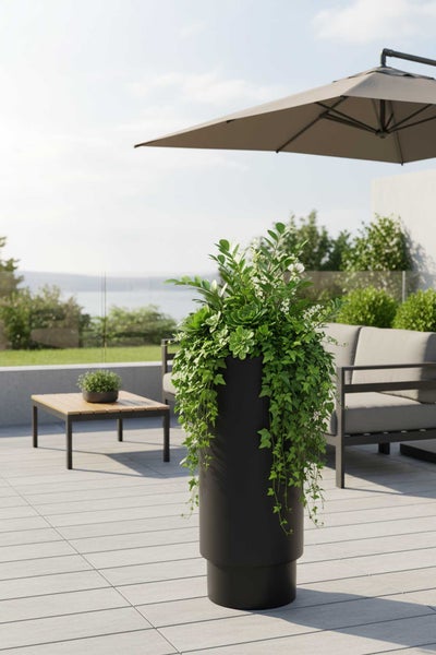 Moderner hoher Pflanzkübel in Schwarz auf einer hellen Terrasse mit Gartenmöbeln und grünen Pflanzen unter einem Sonnenschirm.