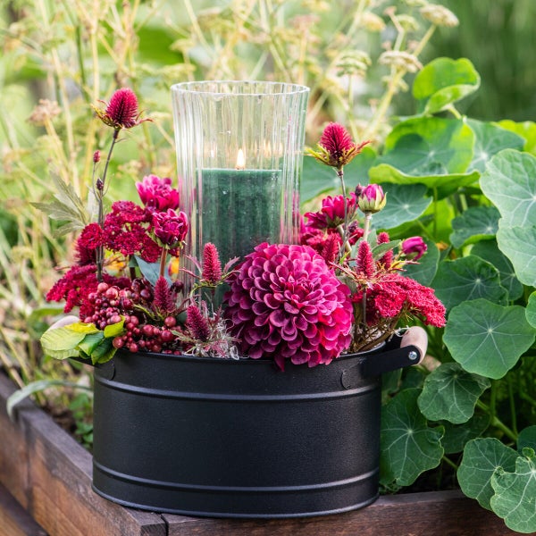 Dekorativer schwarzer Metalltopf mit Henkeln, gefüllt mit Dahlien, Beeren und einem brennenden Windlicht in einer gläsernen Vase im Garten.