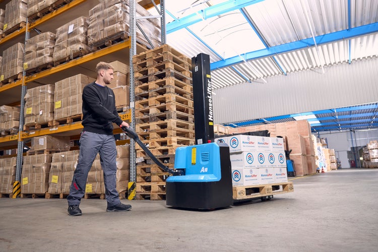 Ein Mitarbeiter bedient einen elektrischen Hochhubwagen von HanseLifter in einem organisierten Lager mit Hochregalen und Holzpaletten.