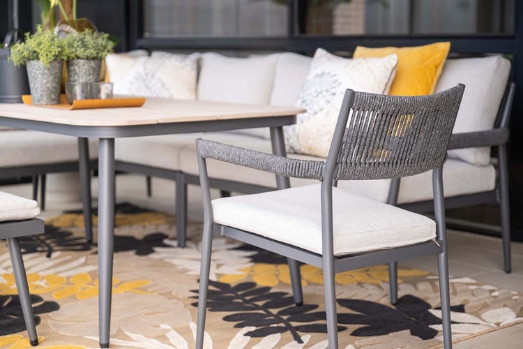 Modernes Garten-Essset mit Armlehnstuhl aus Seilgeflecht, Esstisch mit Holzplatte und Lounge-Sofa mit Kissen auf einem gemusterten Außenteppich.