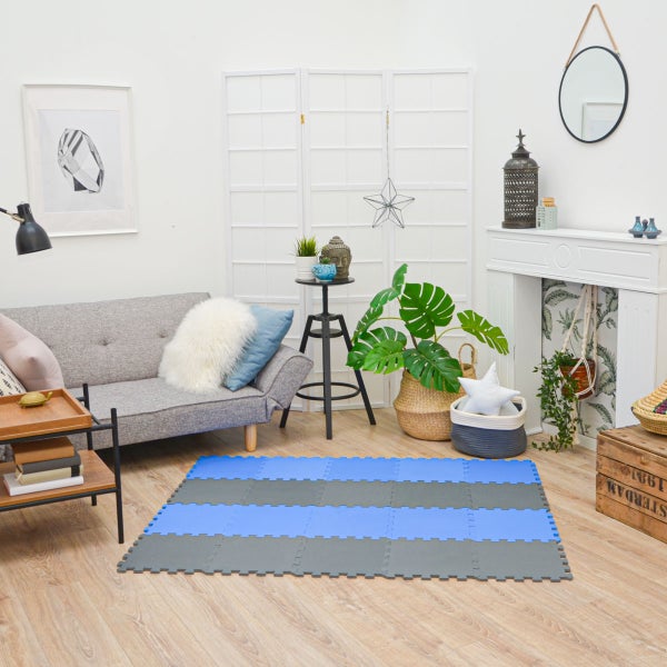Ineinandergreifende blaue und graue Puzzlematten auf Holzboden in einem hellen Wohnzimmer mit grauem Sofa und Pflanzen.