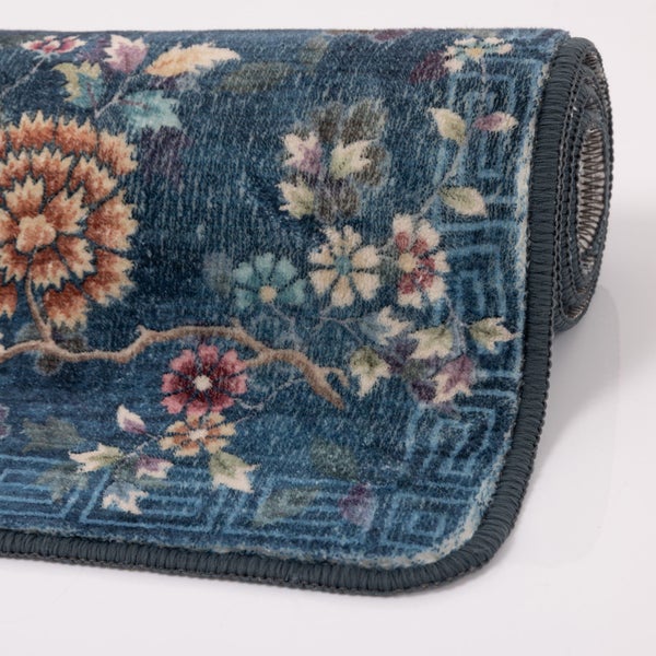 Teilweise aufgerollter blauer Teppich mit Blumenmuster und geometrischer Bordüre, weiche Textur und eingefasste Kanten.